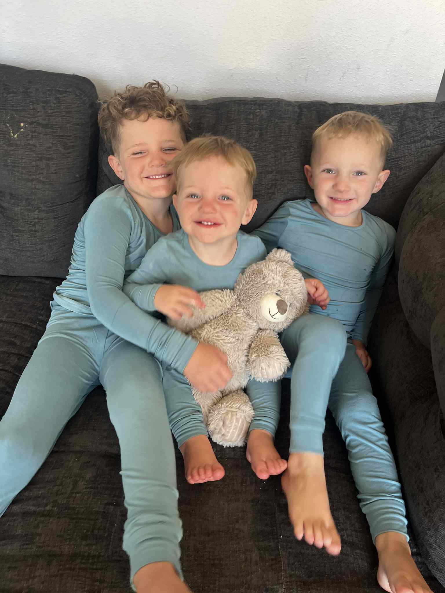 Three children in matching outfits sitting on a couch with a teddy bear.