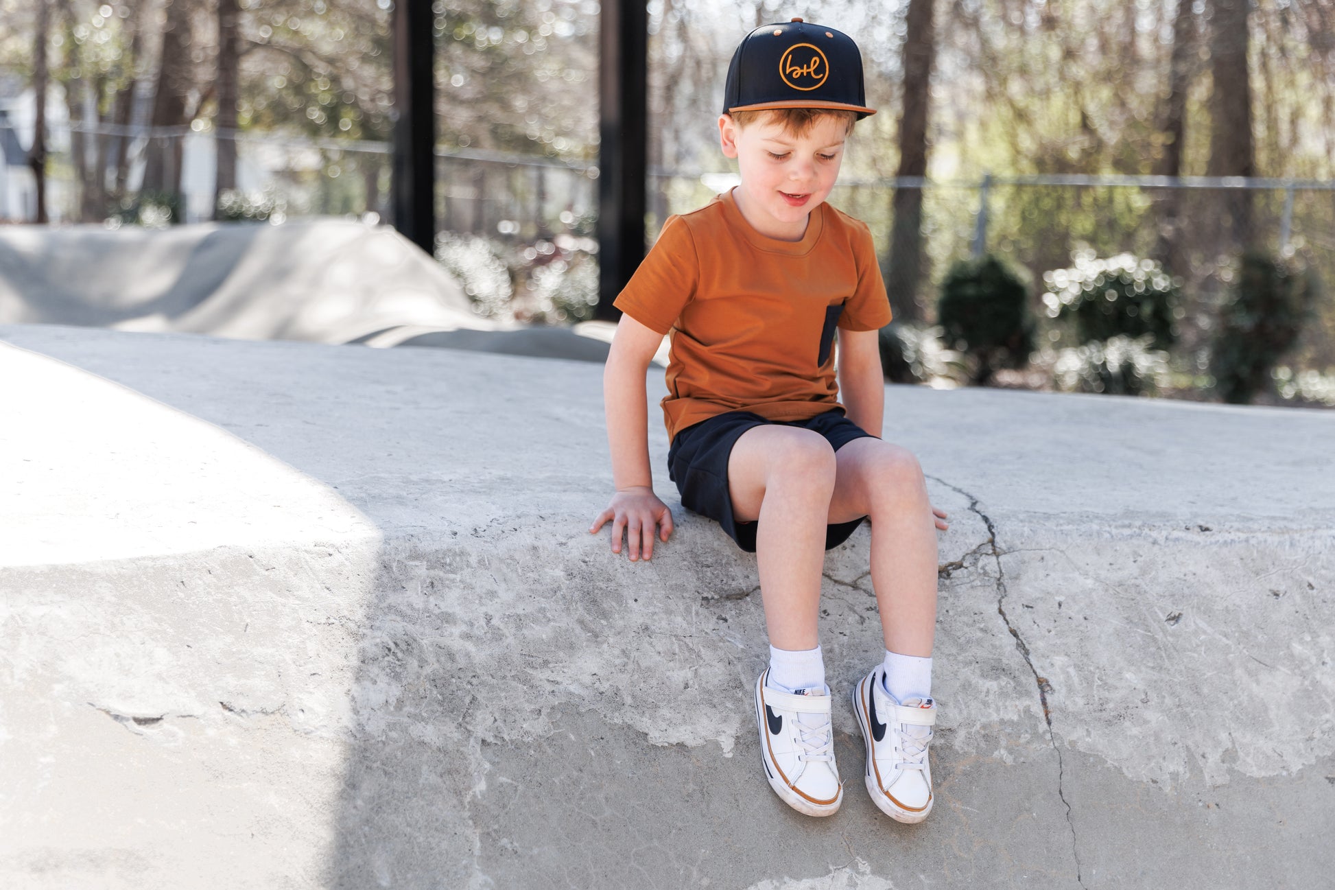 Young boy sitting on a concrete ledge wearing a cap and casual clothing.