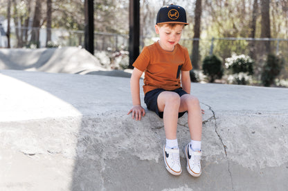 Young boy sitting on a concrete ledge wearing a cap and casual clothing.