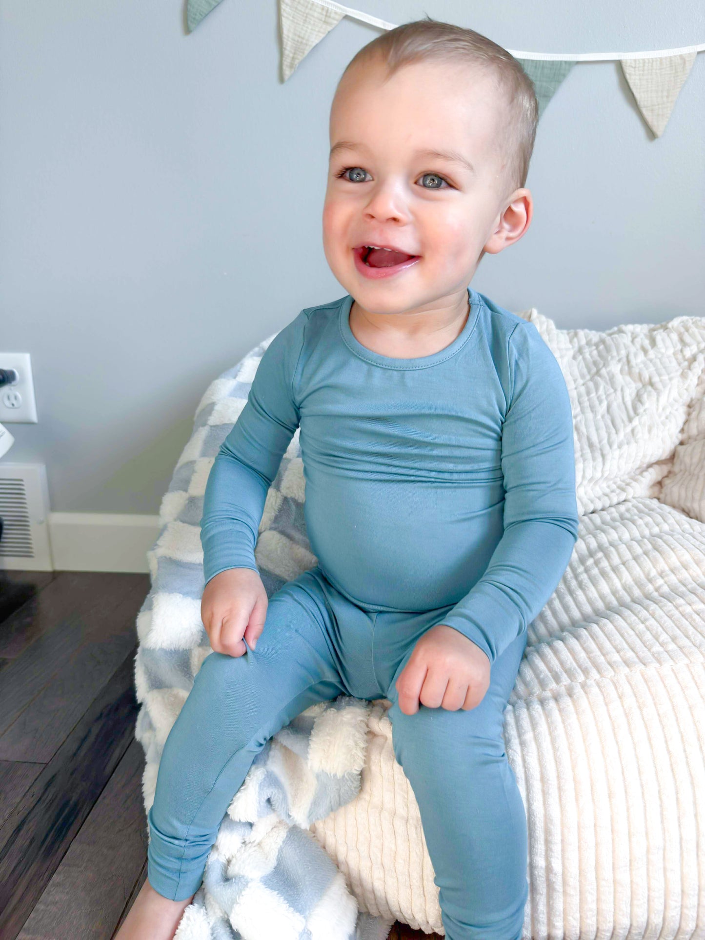 Baby wearing a blue onesie sitting on a bed with a light blue wall in the background