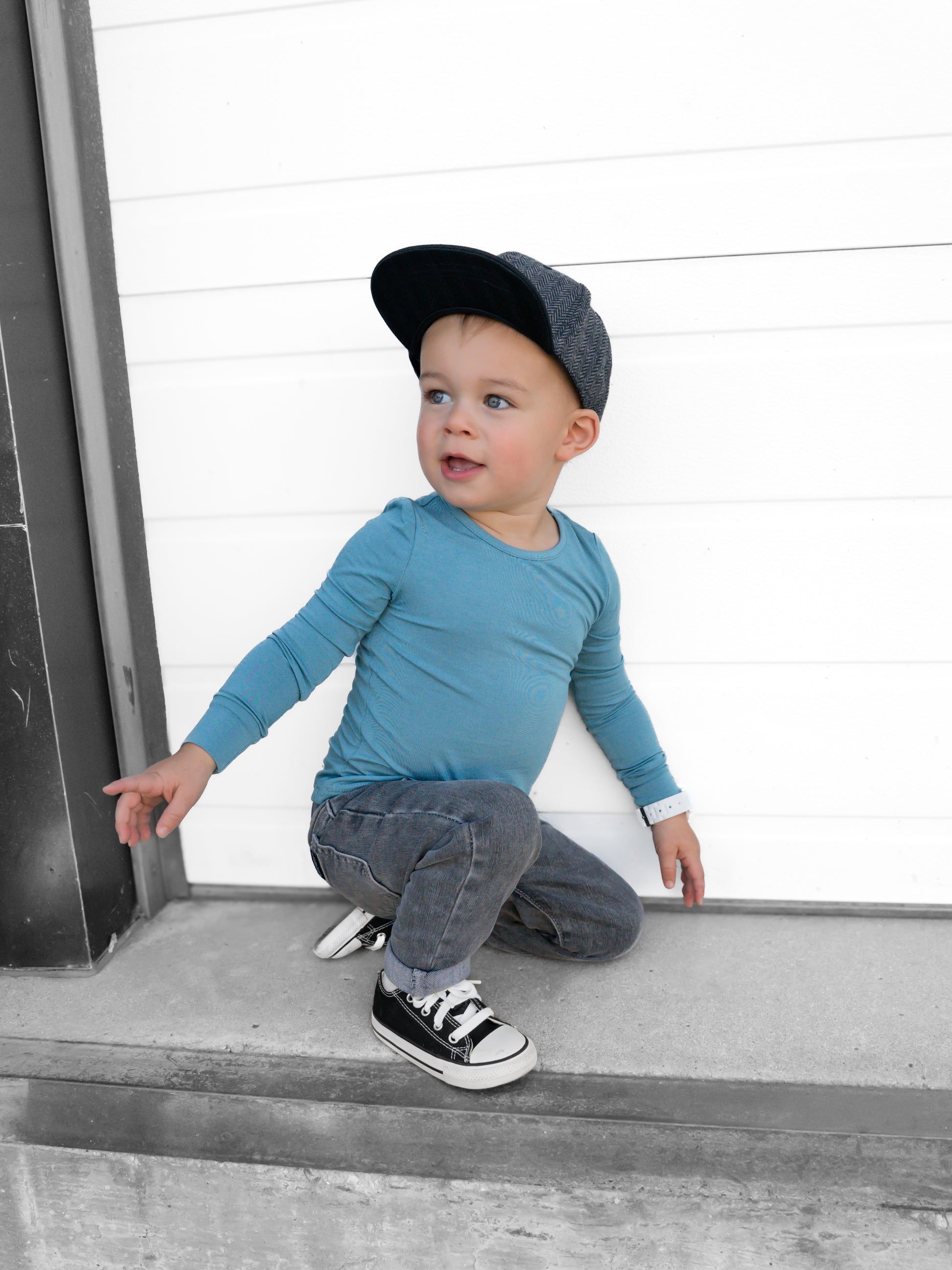 Child wearing a blue shirt and black cap sitting on a concrete ledge with a white wall background