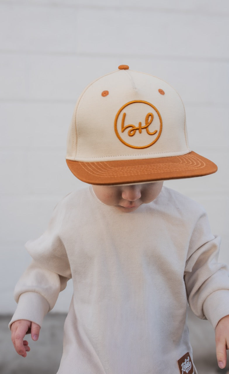 Child wearing a cream cap with brown brim and embroidered logo, standing against a plain background