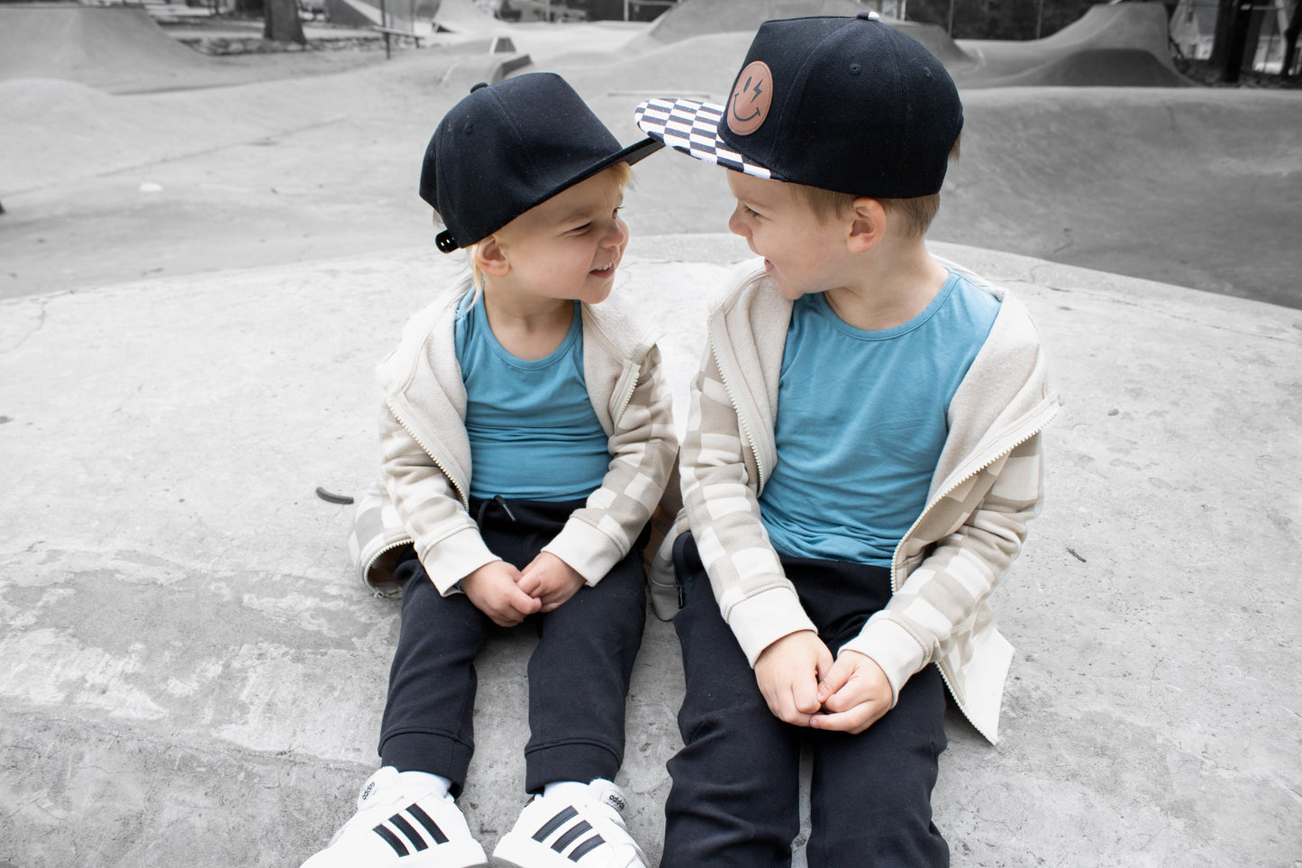 Two children sitting on a concrete surface wearing matching outfits with caps.