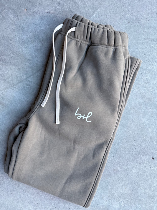 A pair of folded taupe sweatpants with white drawstrings and a white embroidered script logo on the thigh, laid flat on a gray concrete surface.