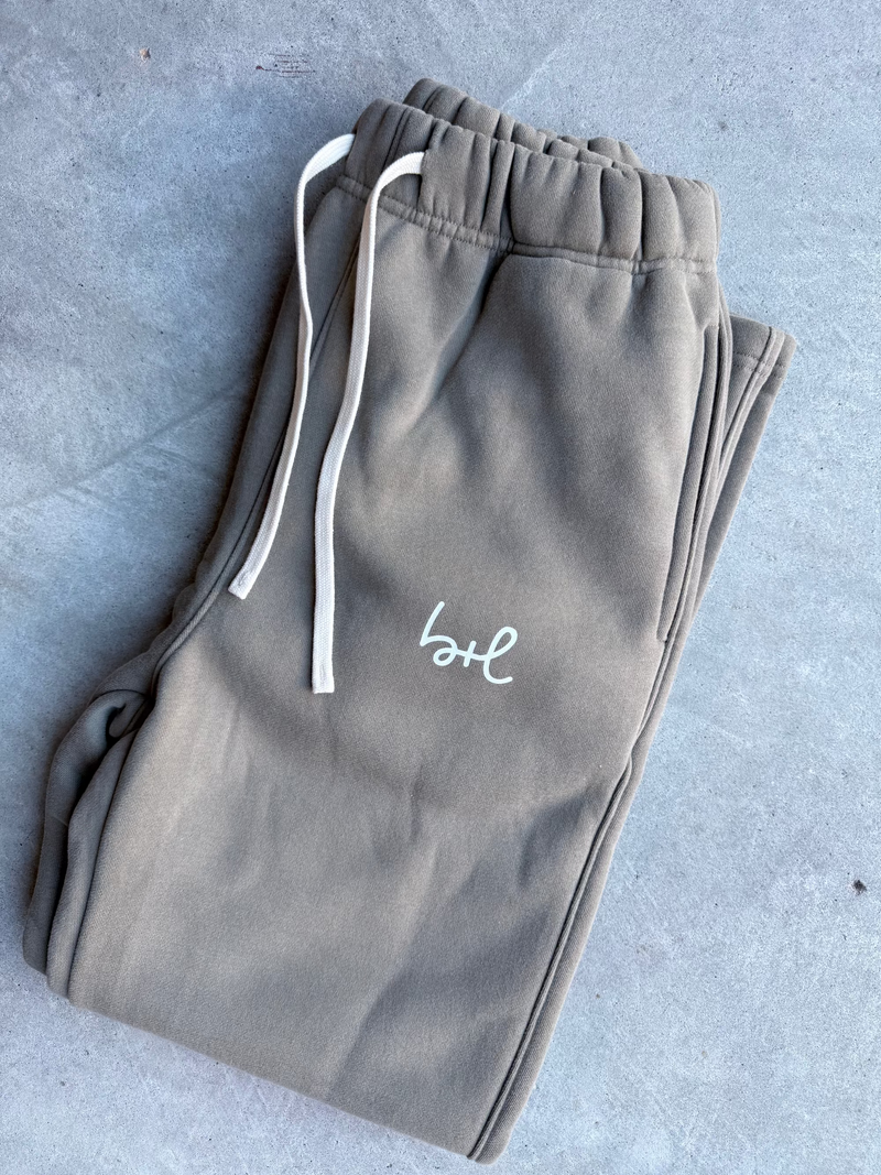 A pair of folded taupe sweatpants with white drawstrings and a white embroidered script logo on the thigh, laid flat on a gray concrete surface.