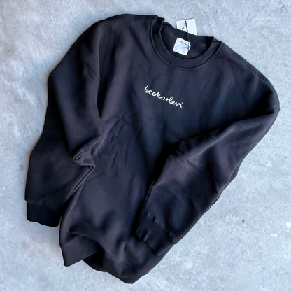 A black crewneck sweatshirt with a small white embroidered script logo on the chest, laid flat on a textured gray concrete surface.