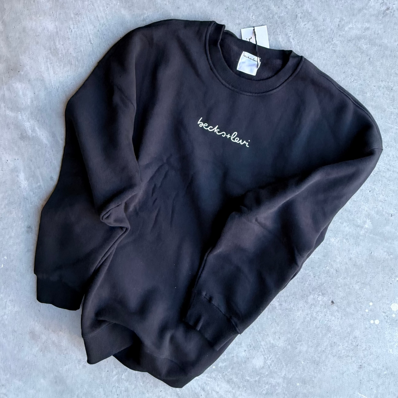 A black crewneck sweatshirt with a small white embroidered script logo on the chest, laid flat on a textured gray concrete surface.